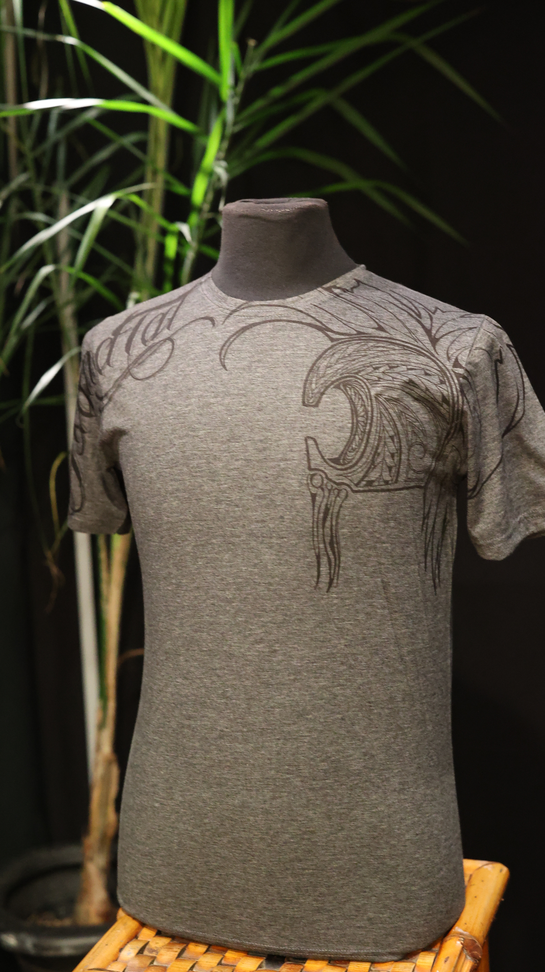 Front angled view of a dark Heather Gray shirt with sweeping Polynesian linework across the shoulders.