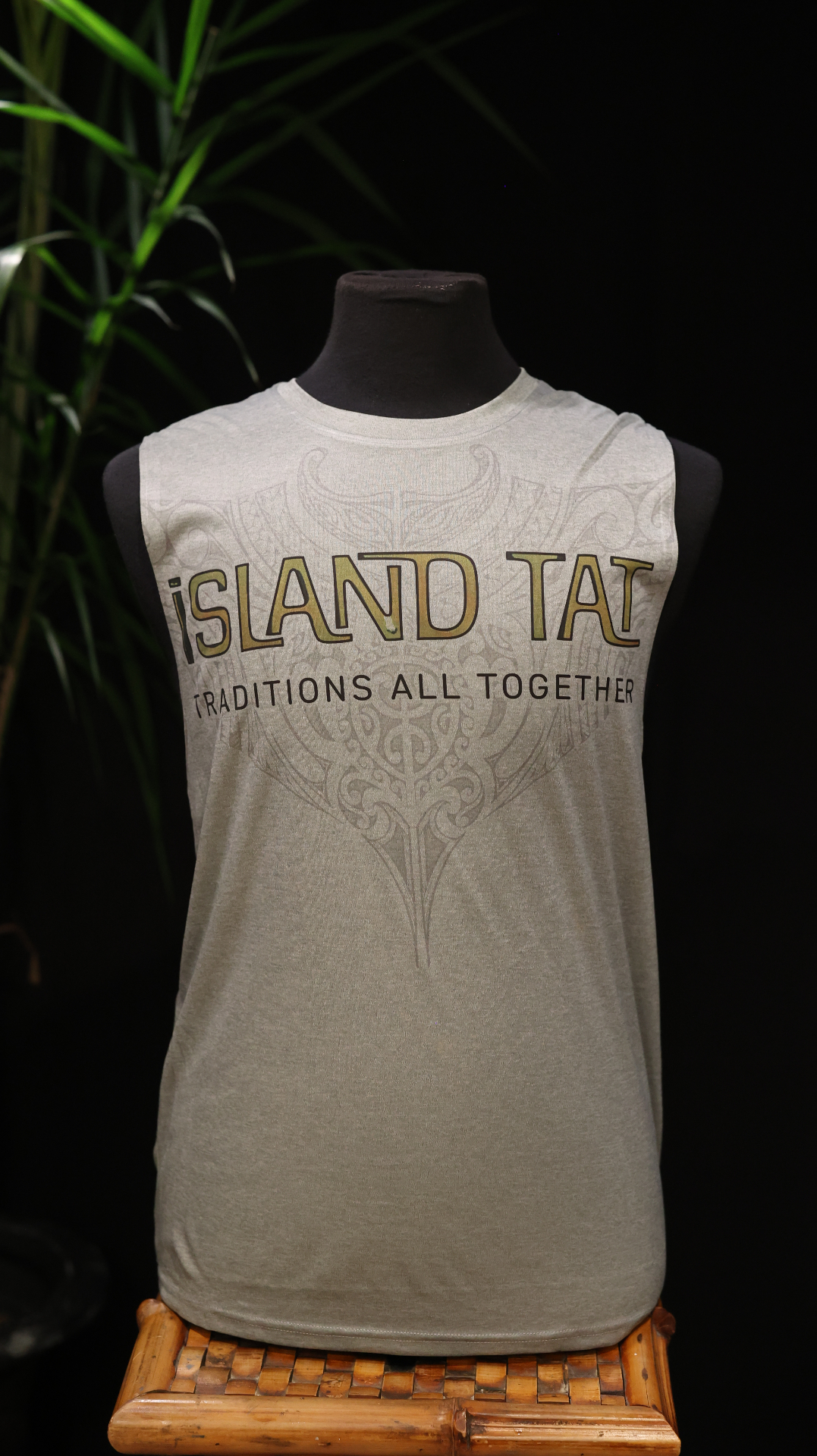 Front view of a natural sleeveless shirt with Island TAT lettering and faint ornamental art behind it.